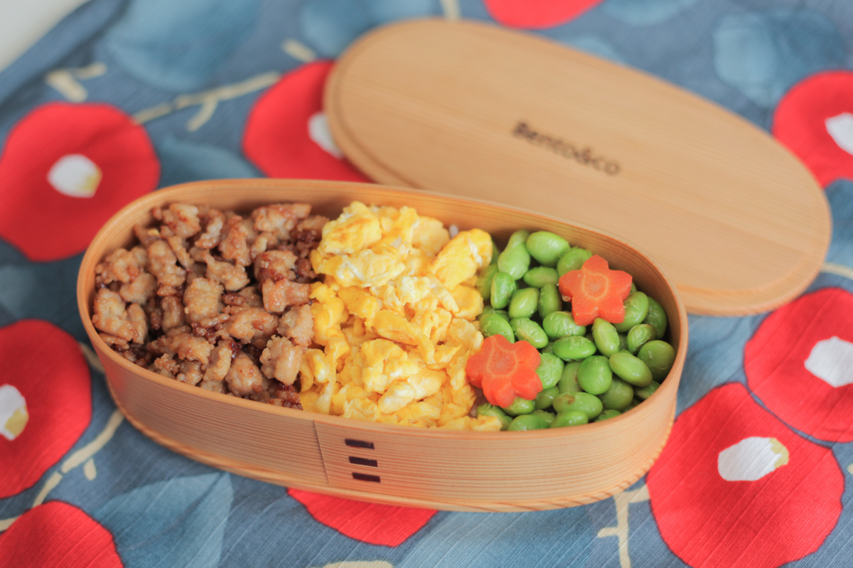 Wooden oval bento box filled with minced meat, eggs and edamame opened on a colorful furoshiki