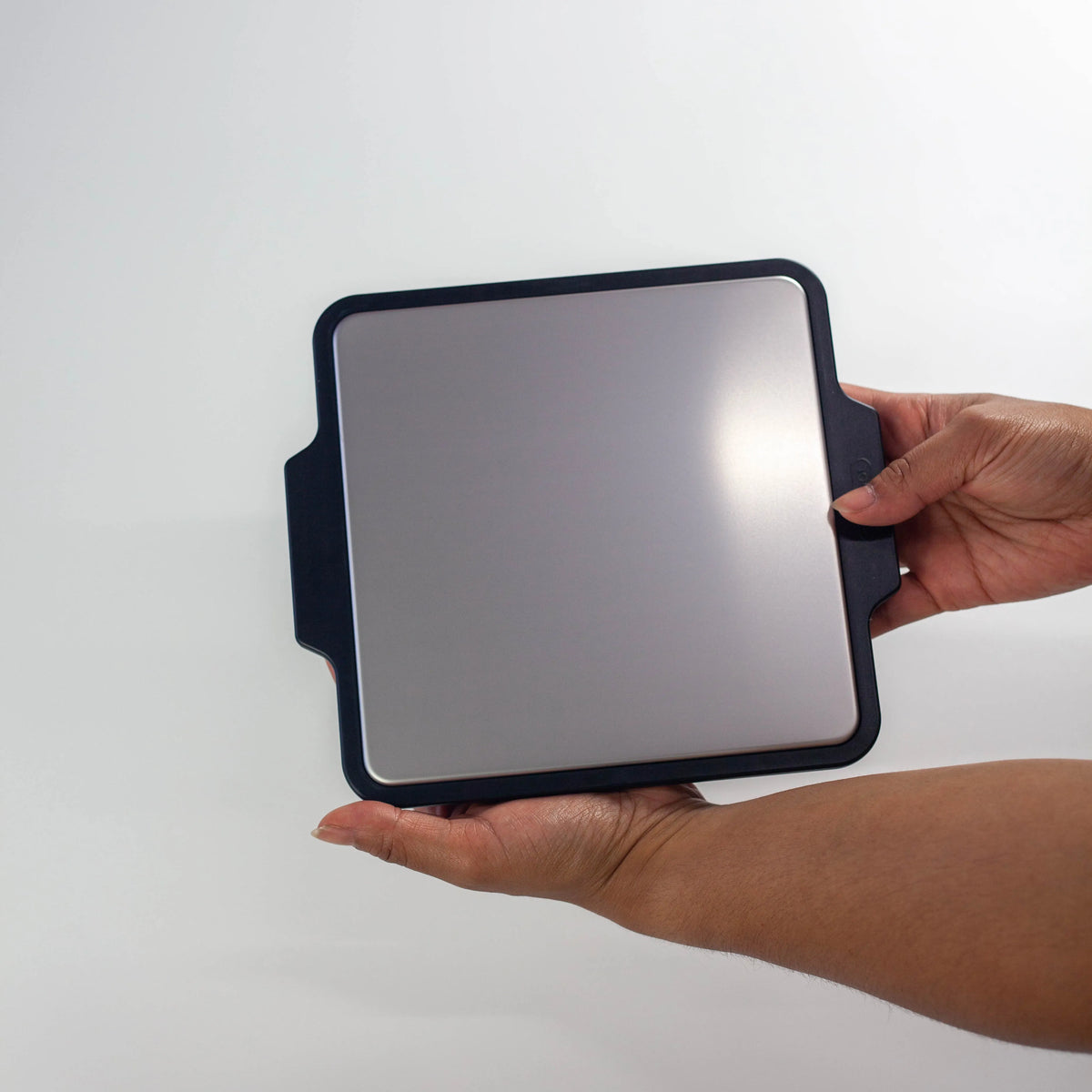 Two hands holding the Gel-Cool Rapid Cooling Plate by its black silicone side handles, showing the size and grip of the aluminum cooling surface, on white background