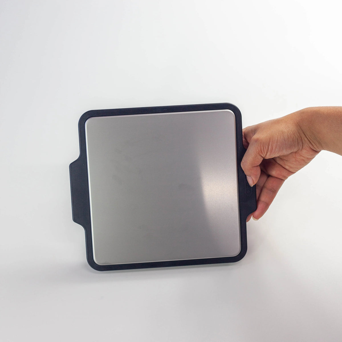 one hand holding the Gel-Cool Rapid Cooling Plate by its black silicone side handles, showing the size and grip of the aluminum cooling surface, on white background