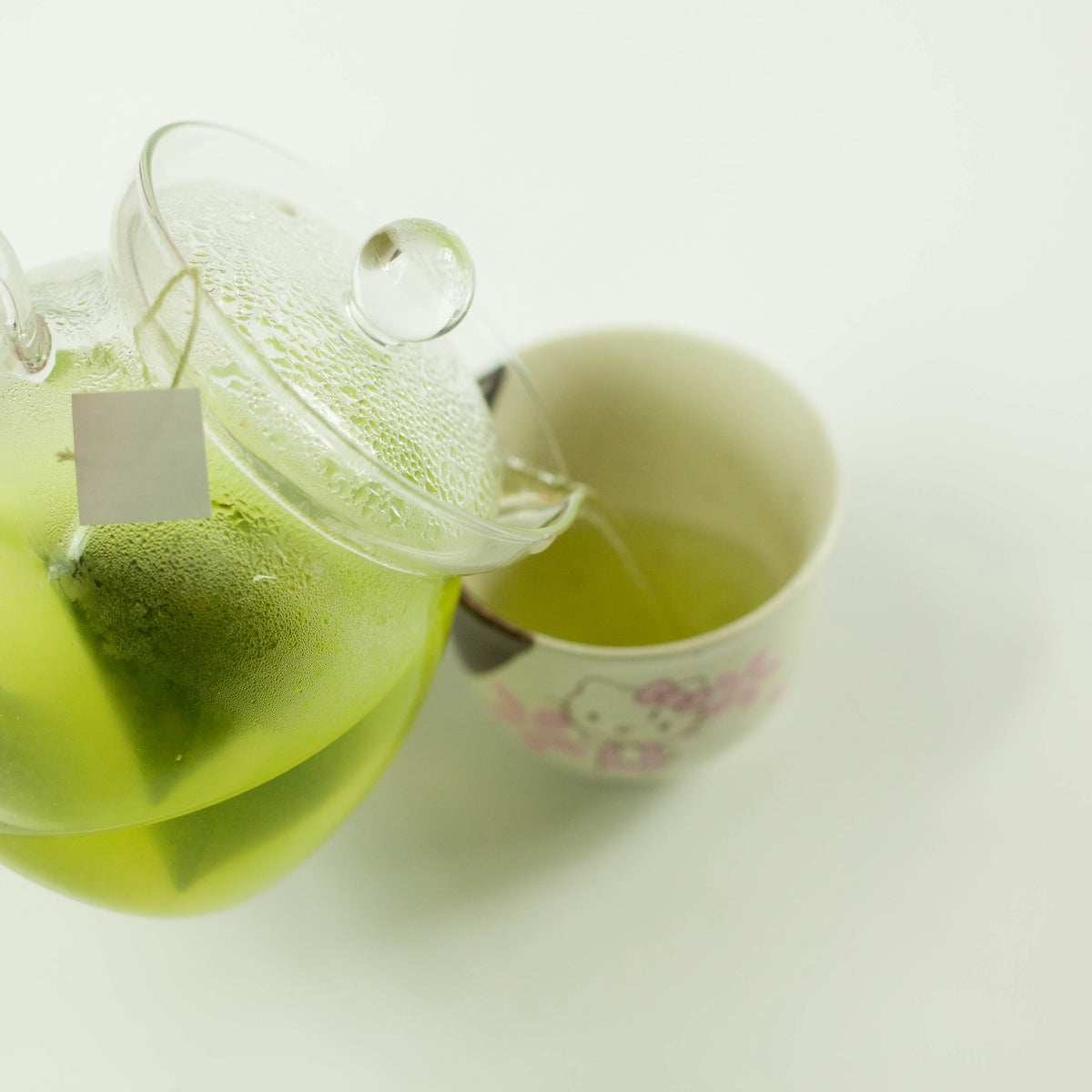 Close-up of cold brew matcha green tea bag steeping in a glass teapot, pouring vivid green tea into a floral ceramic yunomi cup on white background
