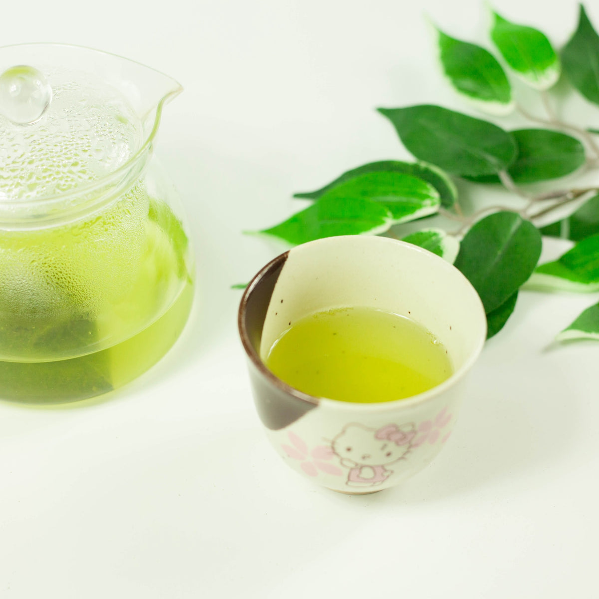 Vibrant green Japanese matcha green tea brewed hot in a glass teapot and poured into a floral ceramic yunomi cup, with fresh green leaves on white background