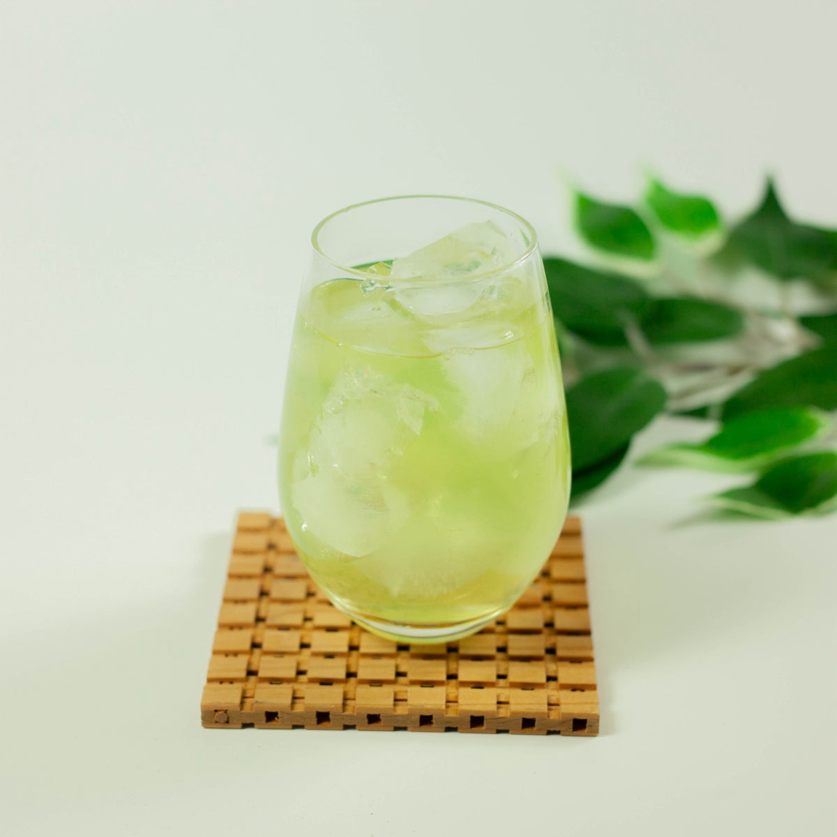 Glass of cold brew matcha green tea over ice on a wooden lattice coaster, with fresh green leaves in the background on white surface
