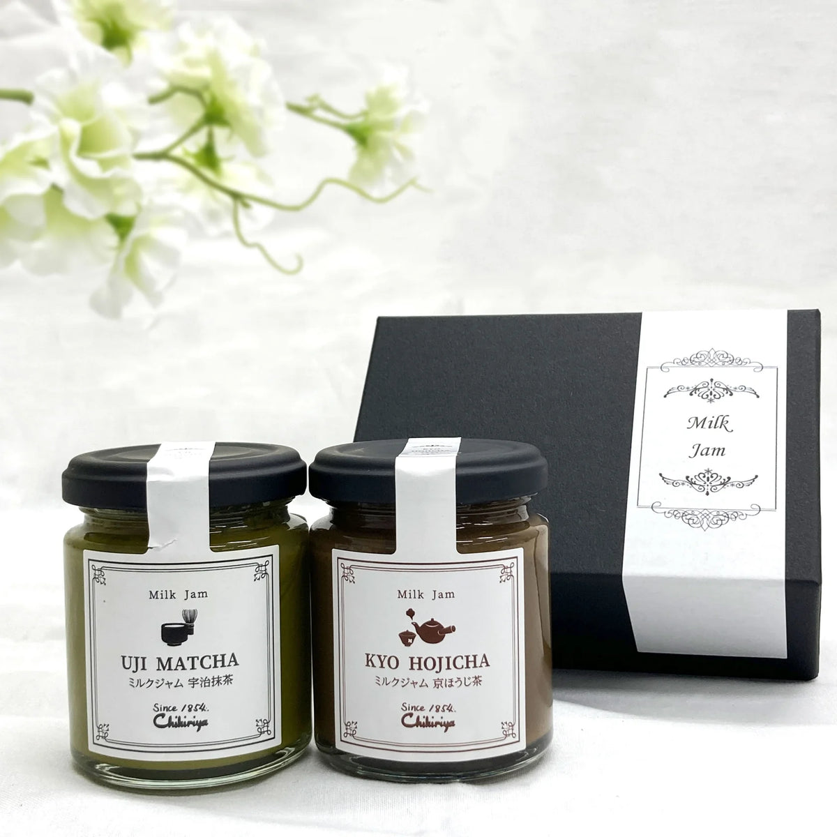 Chikiriya Hojicha and Uji Matcha Milk Jam Gift Set – two glass jars in a white lace-lined wicker basket with white flowers in the background; green Uji Matcha jar on the left, brown Kyo Hojicha jar on the right