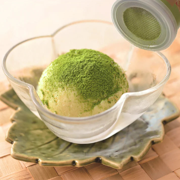 Sifting Chikiriya Uji Matcha Green Tea Powder over a scoop of vanilla ice cream in a glass leaf-shaped bowl on a ceramic plate, using the built-in sifter lid