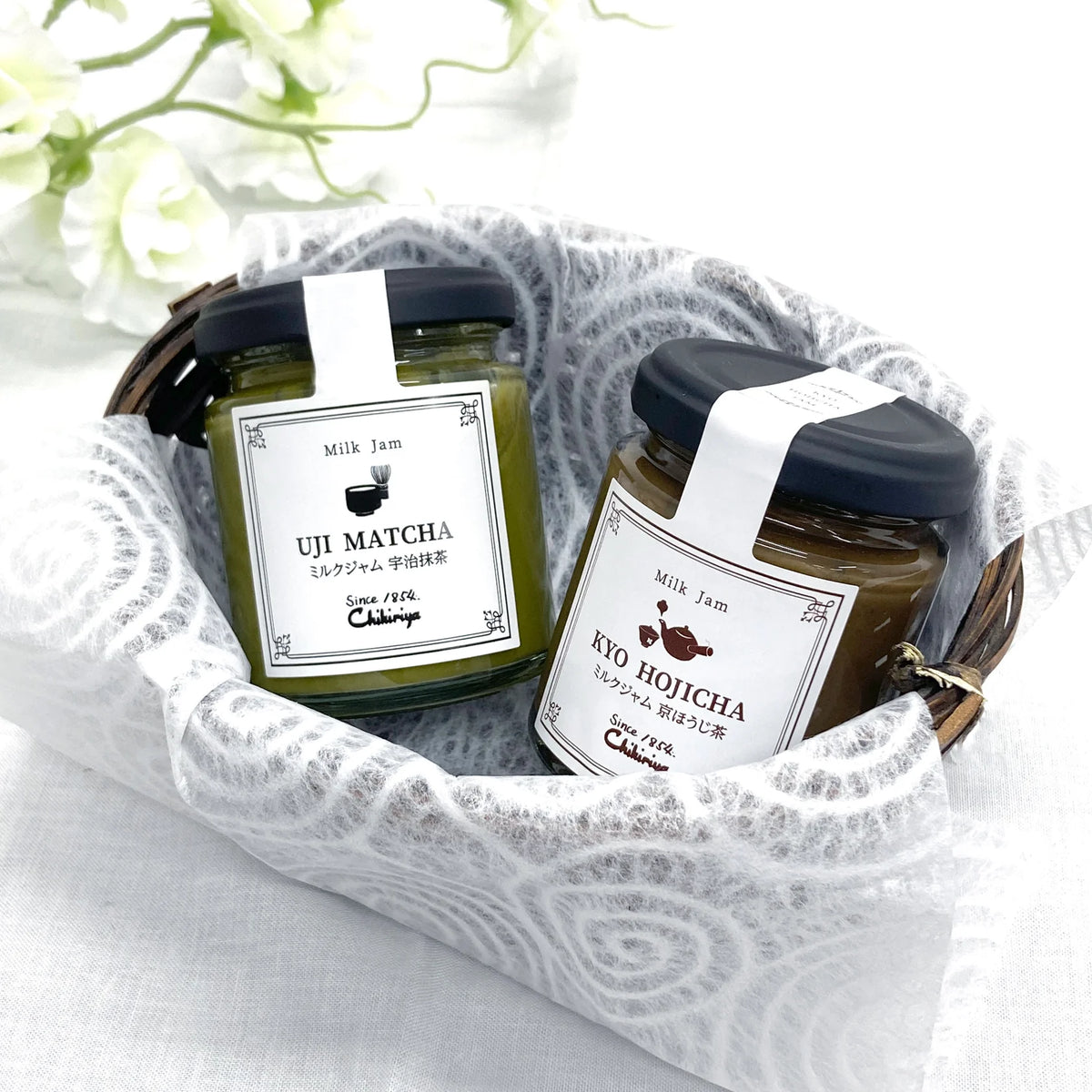 Chikiriya Hojicha and Uji Matcha Milk Jam Gift Set – two glass jars in a white lace-lined wicker basket with white flowers in the background; green Uji Matcha jar on the left, brown Kyo Hojicha jar on the right