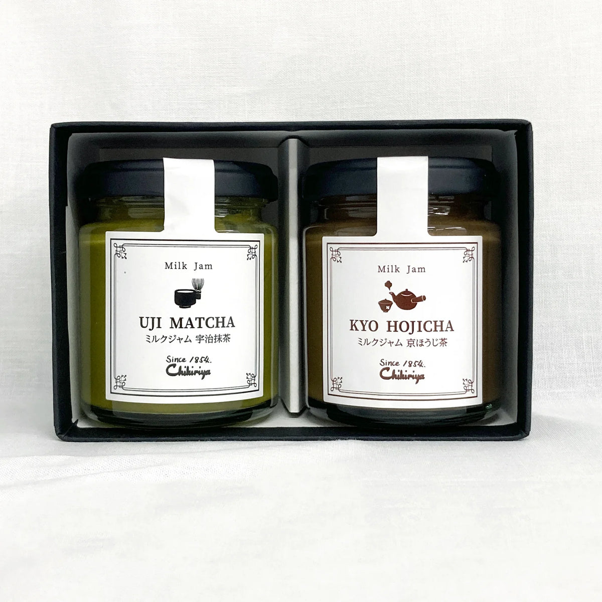 Chikiriya Hojicha and Uji Matcha Milk Jam Gift Set – two glass jars in a white lace-lined wicker basket with white flowers in the background; green Uji Matcha jar on the left, brown Kyo Hojicha jar on the right