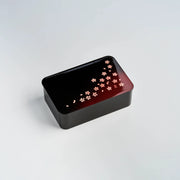 Hakoya Sakura Petals Bento Box closed, black to burgundy gradient lacquer lid with scattered pink cherry blossom pattern, angled view on grey background