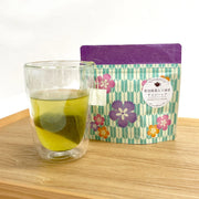 Chikiriya Sencha with Uji Matcha tea bag steeping in a double-walled glass mug showing vivid yellow-green tea, beside the teal geometric floral Japanese-pattern resealable pouch with purple top, on a wooden surface