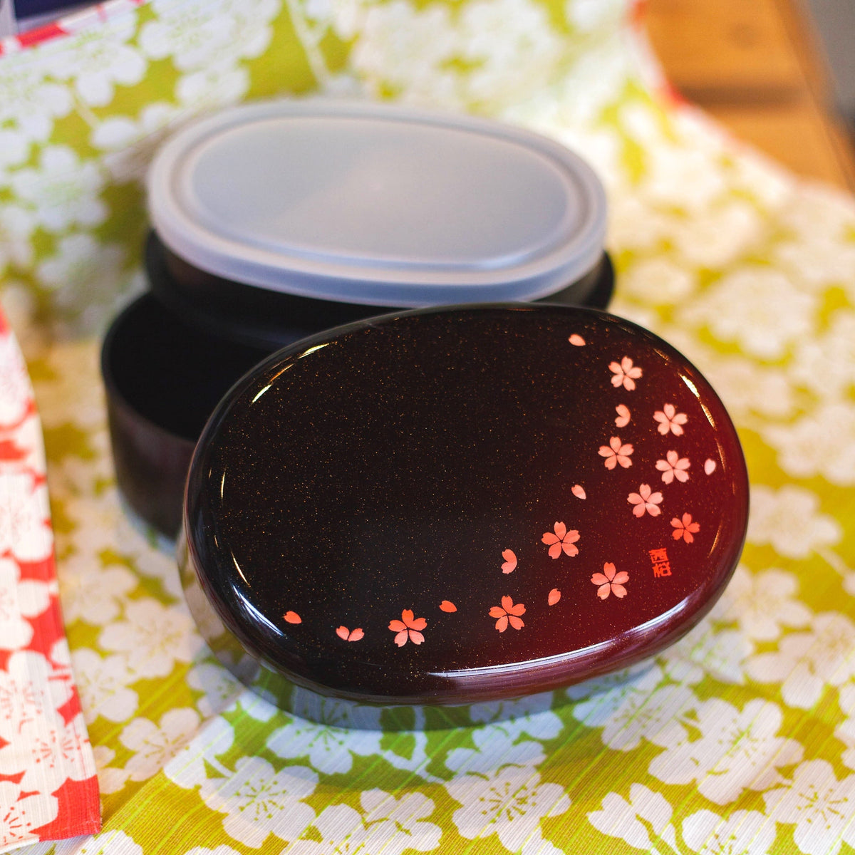 Hakoya Sakura Petals Oval Bento Box closed, black to burgundy lacquer with scattered pink cherry blossom pattern, angled view on grey background