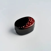 Hakoya Sakura Petals Oval Bento Box closed, black to burgundy lacquer with scattered pink cherry blossom pattern, angled view on grey background
