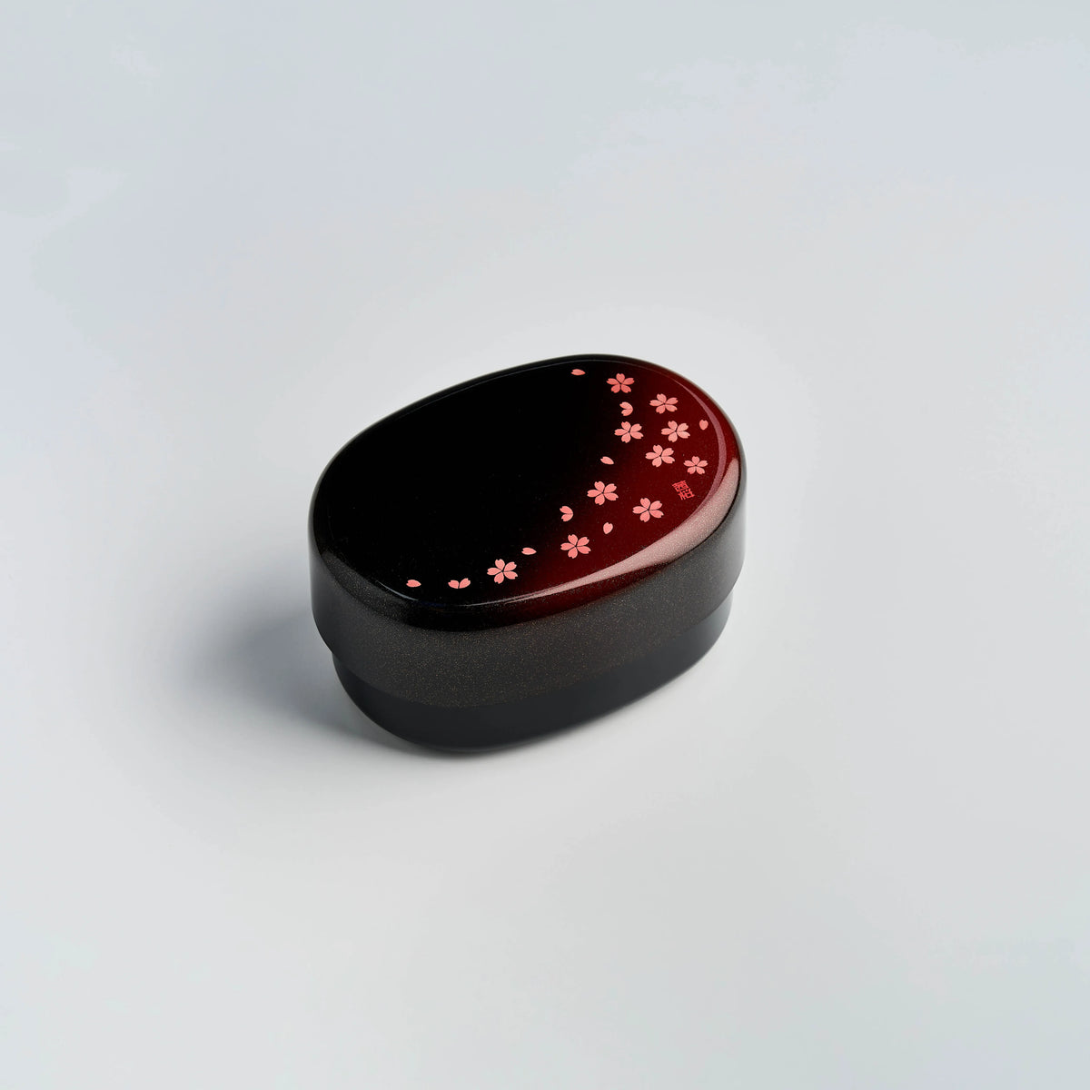 Hakoya Sakura Petals Oval Bento Box closed, black to burgundy lacquer with scattered pink cherry blossom pattern, angled view on grey background