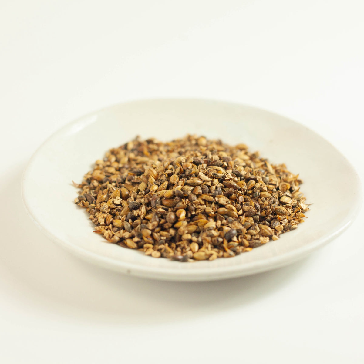 Close-up angled view of roasted barley grains for mugicha tea piled on a white ceramic plate on white background