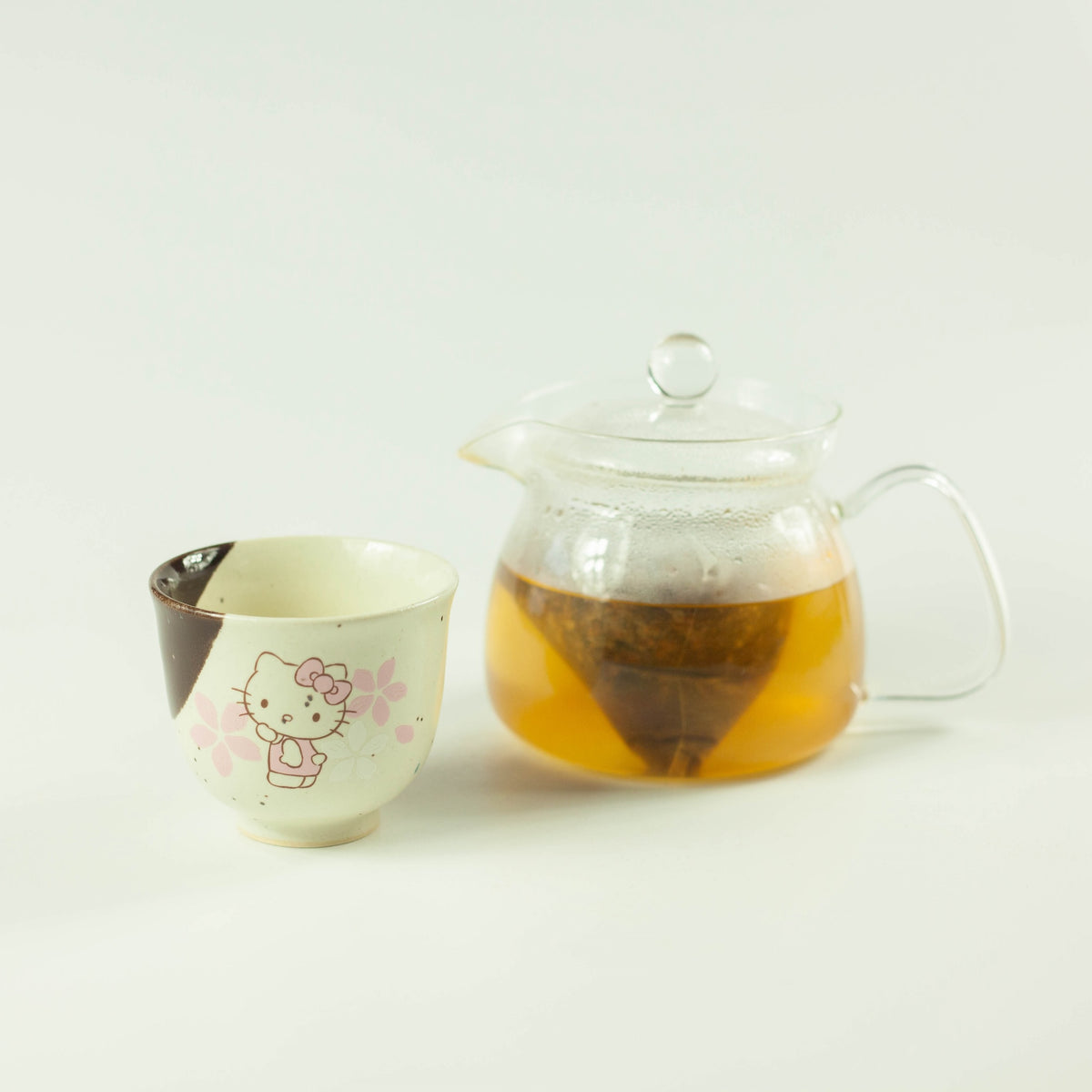 Glass of cold brew mugicha barley tea over ice beside a glass teapot with a tea bag steeping inside, with fresh green leaves on white background