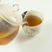 Pouring brewed Japanese mugicha barley tea from a glass teapot into a Hello Kitty ceramic cup on a white background