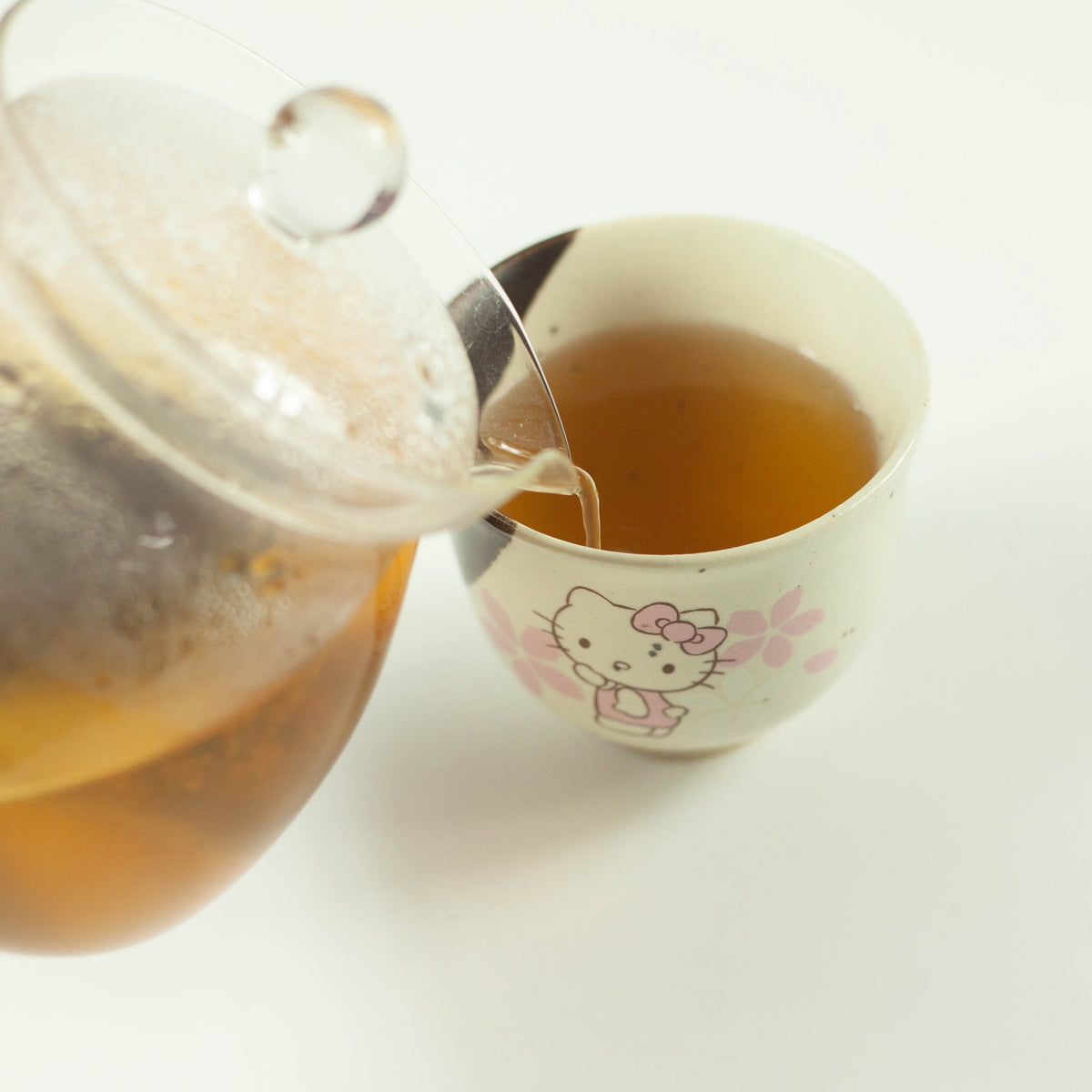 Pouring brewed Japanese mugicha barley tea from a glass teapot into a Hello Kitty ceramic cup on a white background