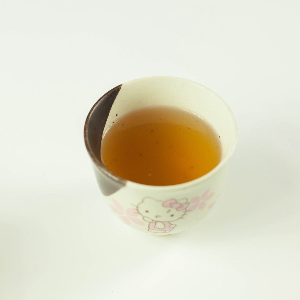 Cup of cold brew mugicha barley tea over ice beside a glass teapot with a tea bag steeping inside, with fresh green leaves on white background