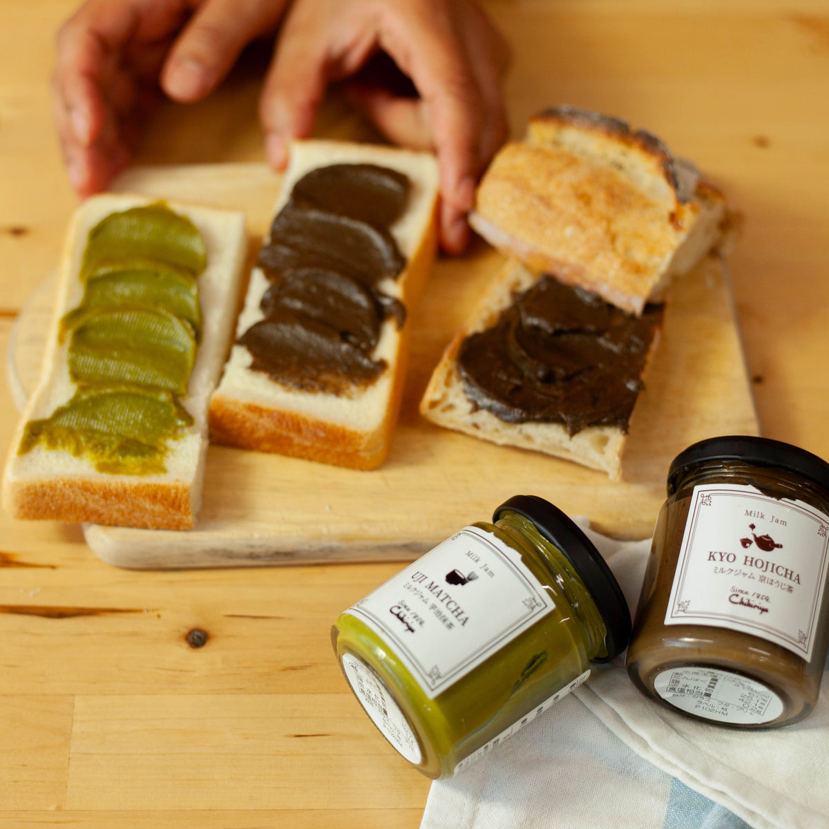 Hands spreading Chikiriya Kyo Hojicha Milk Jam onto a slice of white bread with a purple knife on a wooden cutting board, beside another slice spread with Uji Matcha Milk Jam