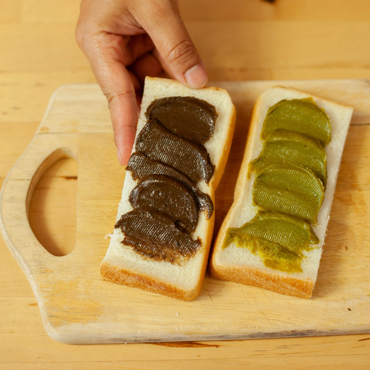Hand picking up a slice of white bread spread with Chikiriya Kyo Hojicha Milk Jam beside a slice spread with Uji Matcha Milk Jam on a wooden cutting board – dark brown hojicha on the left, vivid green matcha on the right