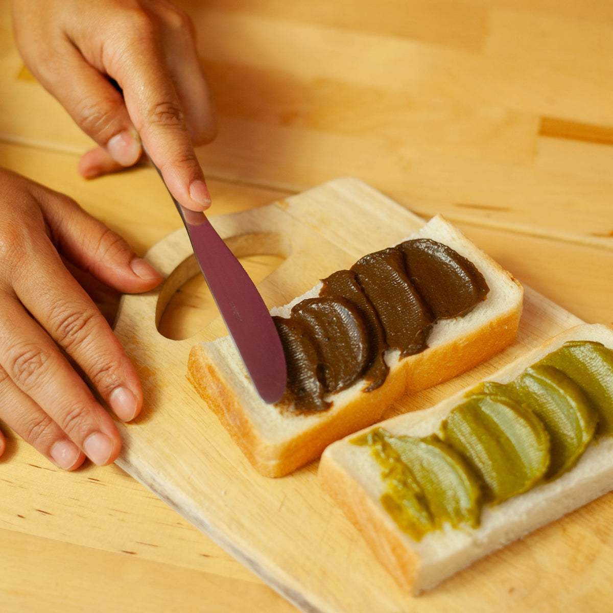 Hands spreading Chikiriya Kyo Hojicha Milk Jam onto a slice of white bread with a purple knife on a wooden cutting board, beside another slice spread with Uji Matcha Milk Jam
