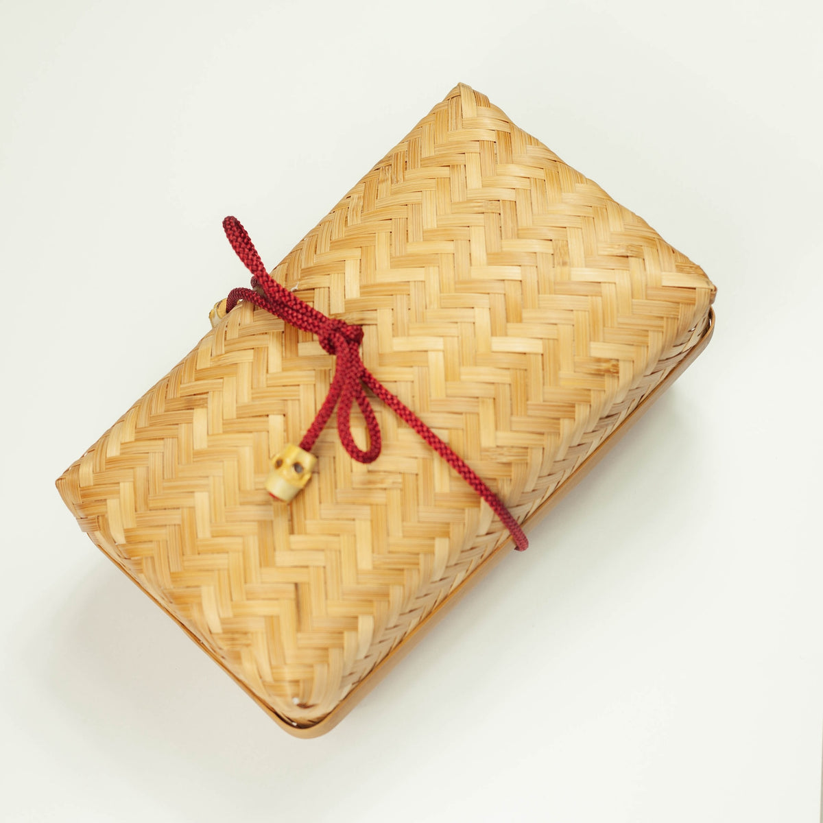 Woven Bamboo Bento Box – honey-brown herringbone woven bamboo with red braided cord and wooden bead clasp, angled view on white background
