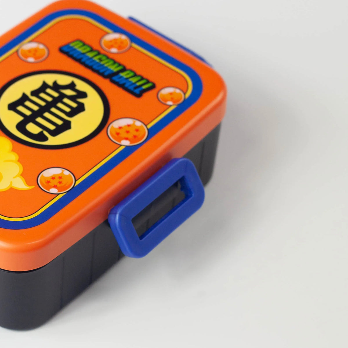 Open Dragon Ball bento box showing a black interior with a divider and an orange lid with a white seal and four blue clips