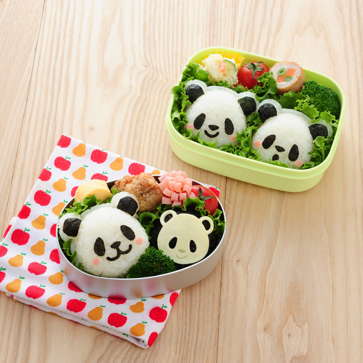 Two bento boxes on a wooden table filled with panda-shaped onigiri made with the Arnest Omusubi Panda Mold Set – a round silver tin with a large panda and a small cheese panda, and a green rectangular box with two panda rice balls, broccoli, cherry tomatoes, and rolled egg