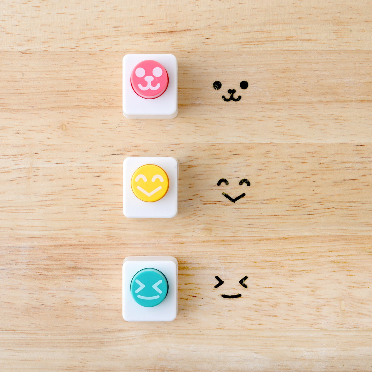 Three Arnest Nico Nico Nori Puncher Mini tools on a wooden surface – pink bear-face punch, yellow happy-face punch, and teal winking-face punch – each shown beside their corresponding nori cutout impression
