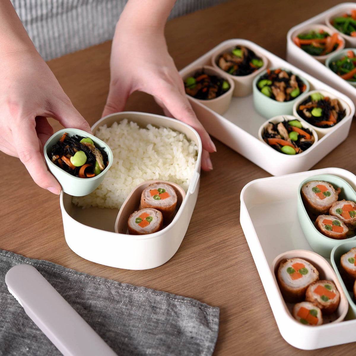 Hands placing silicone cup with hijiki salad into Marumi bento tray filled with rice and meat rolls, multiple trays in background