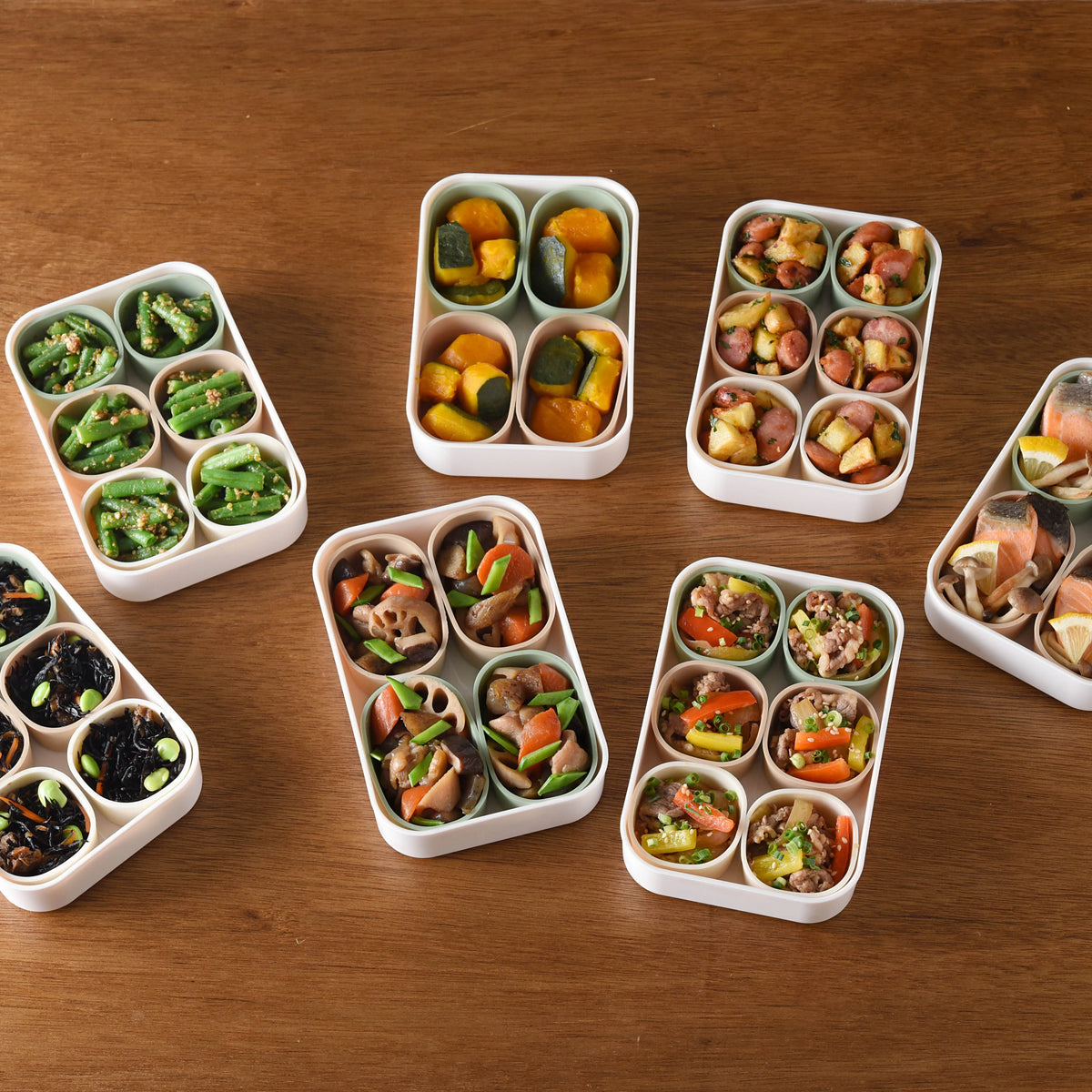Multiple Marumi Silicone Cup Sets Round filled with Japanese meal-prep side dishes – green beans, kabocha, sausage, lotus root stew, ground meat stir-fry, and hijiki, overhead view on wooden table