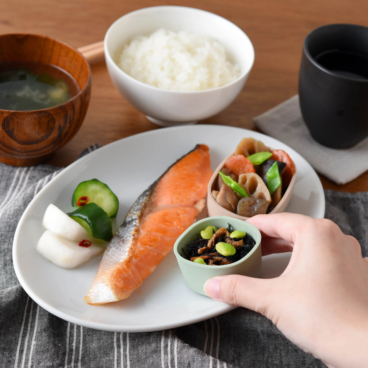 Hand placing Marumi silicone cup with hijiki salad onto a Japanese breakfast plate with grilled salmon, pickled vegetables, rice, miso soup, and tea