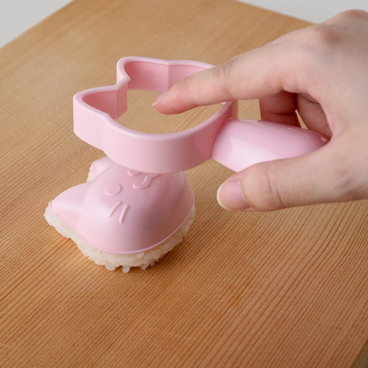 Hands filling pink cat-shaped rice mold from Neko Kao Onigiri Set with white rice on wooden surface