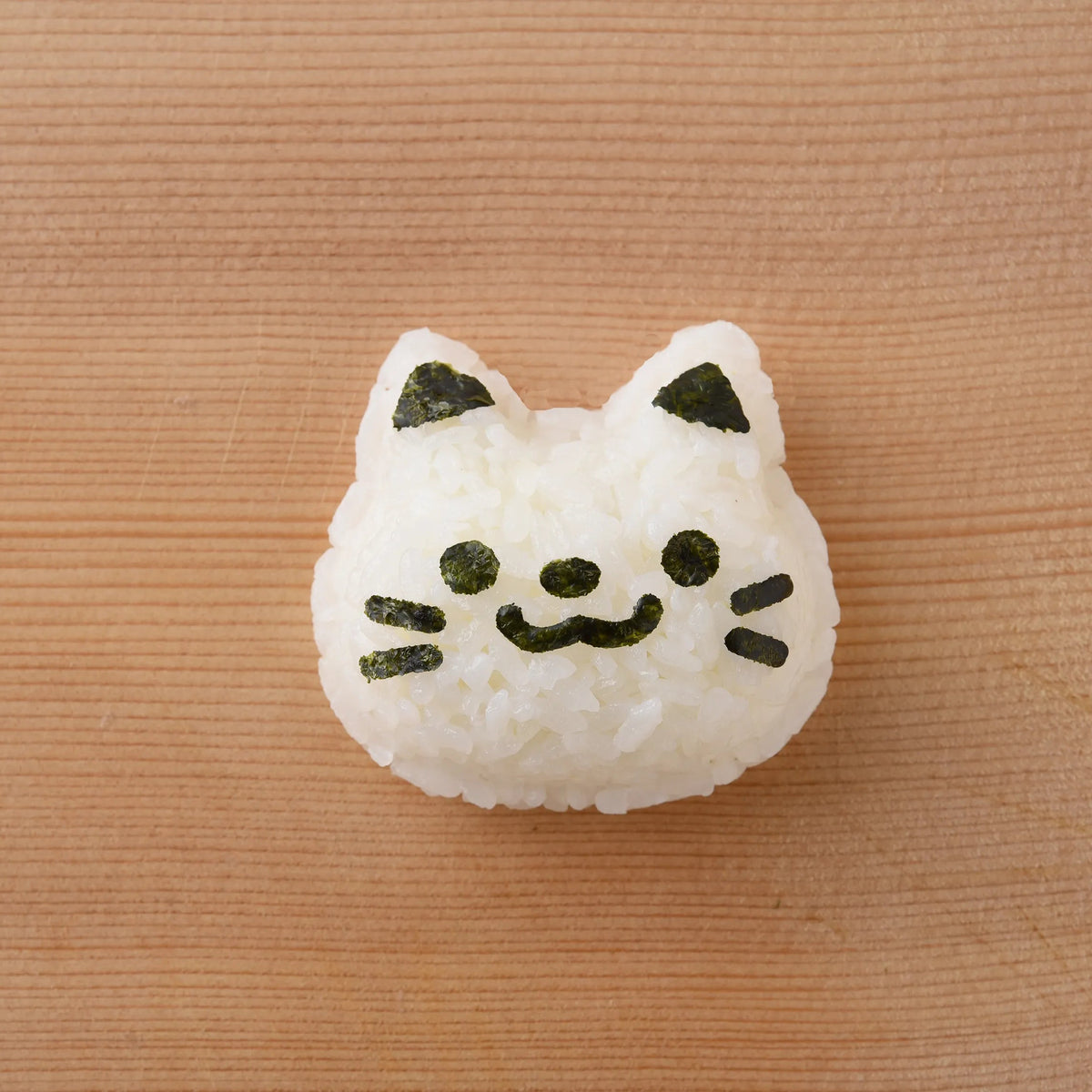 Finished cat face onigiri made with Neko Kao Onigiri Set – white rice shaped into a smiling cat with nori eyes, nose, whiskers, and ear accents, on a wooden surface