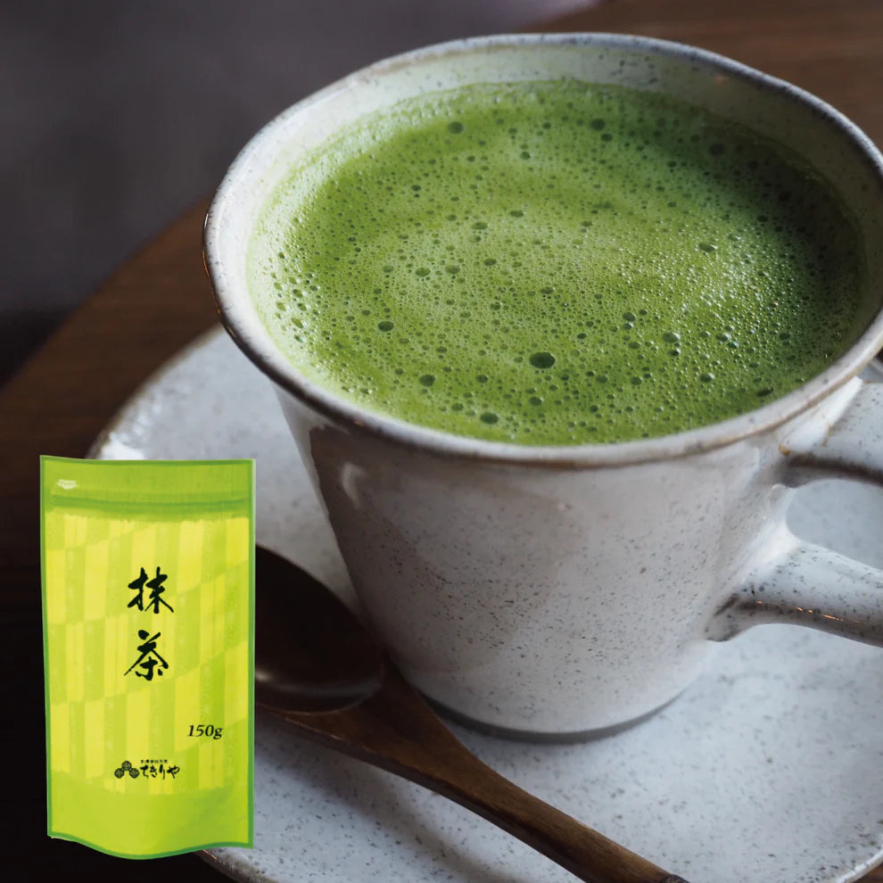 Frothy matcha latte in a white ceramic mug on a saucer with a wooden spoon, alongside the Chikiriya Shizuoka Matcha Powder 150g green pouch