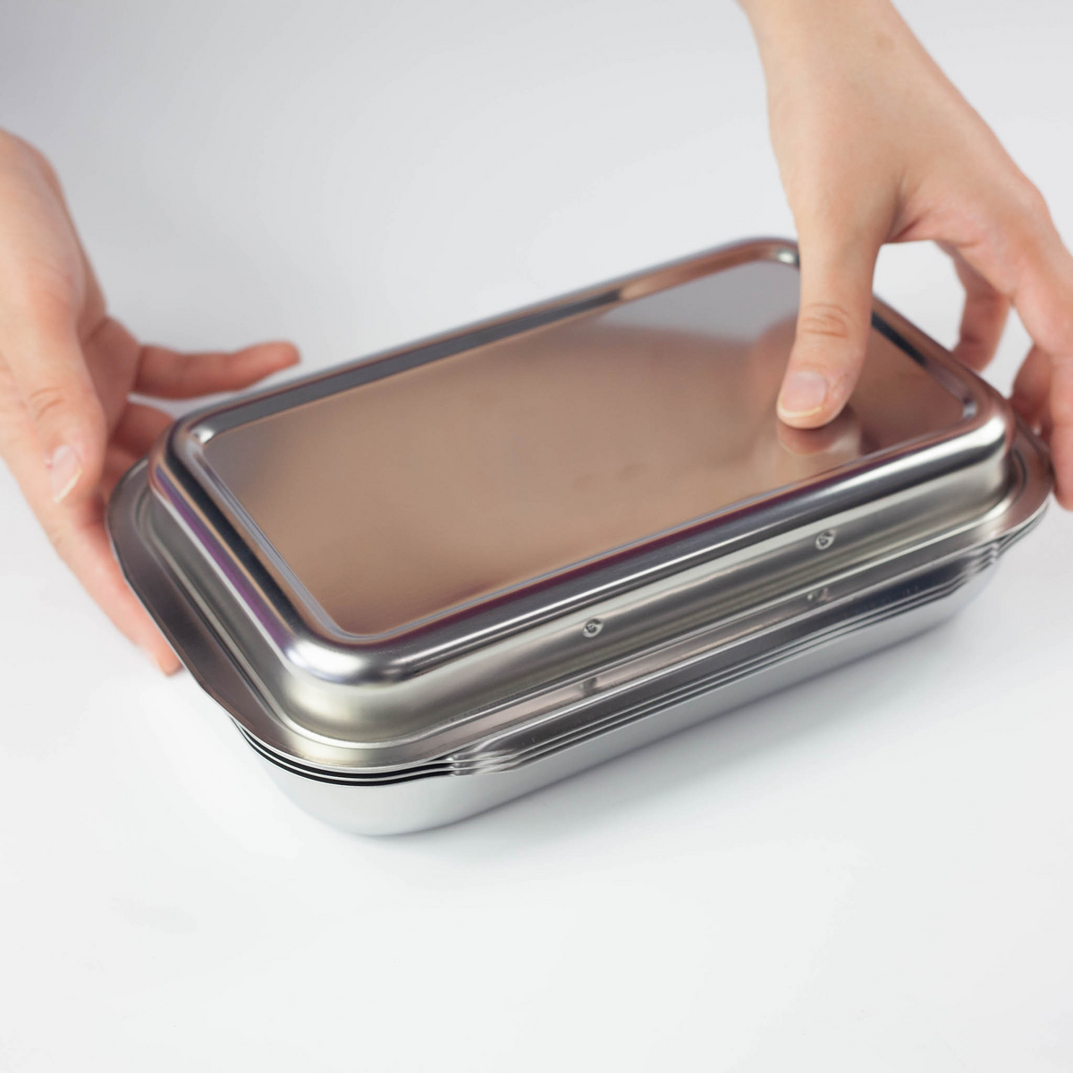 Hands closing the lid of Compact Vat Set – stainless steel rectangular tray with fitted lid, angled view on white background
