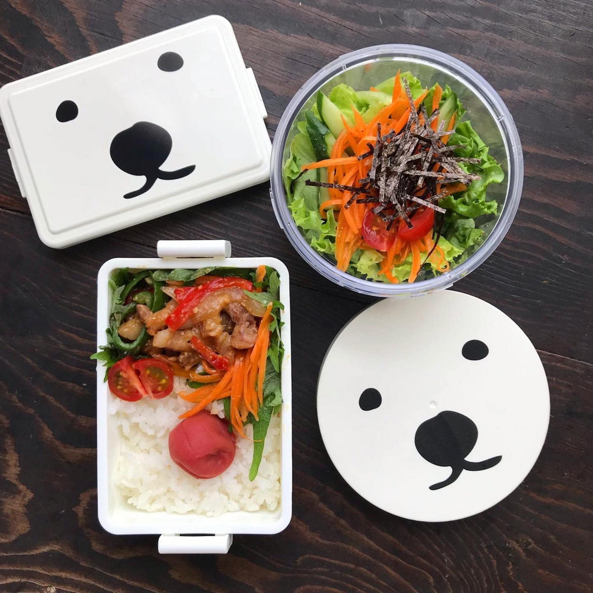 Overhead view of the Gel-Cooma series on a dark wooden table – Gel-Cooma Bento Box Medium 400ml open with white rice, stir-fried meat and vegetables, cherry tomatoes, and a pickled plum, beside the Gel-Cooma Round Salad Case filled with green salad, shredded carrots, and nori, with both bear-face lids placed nearby