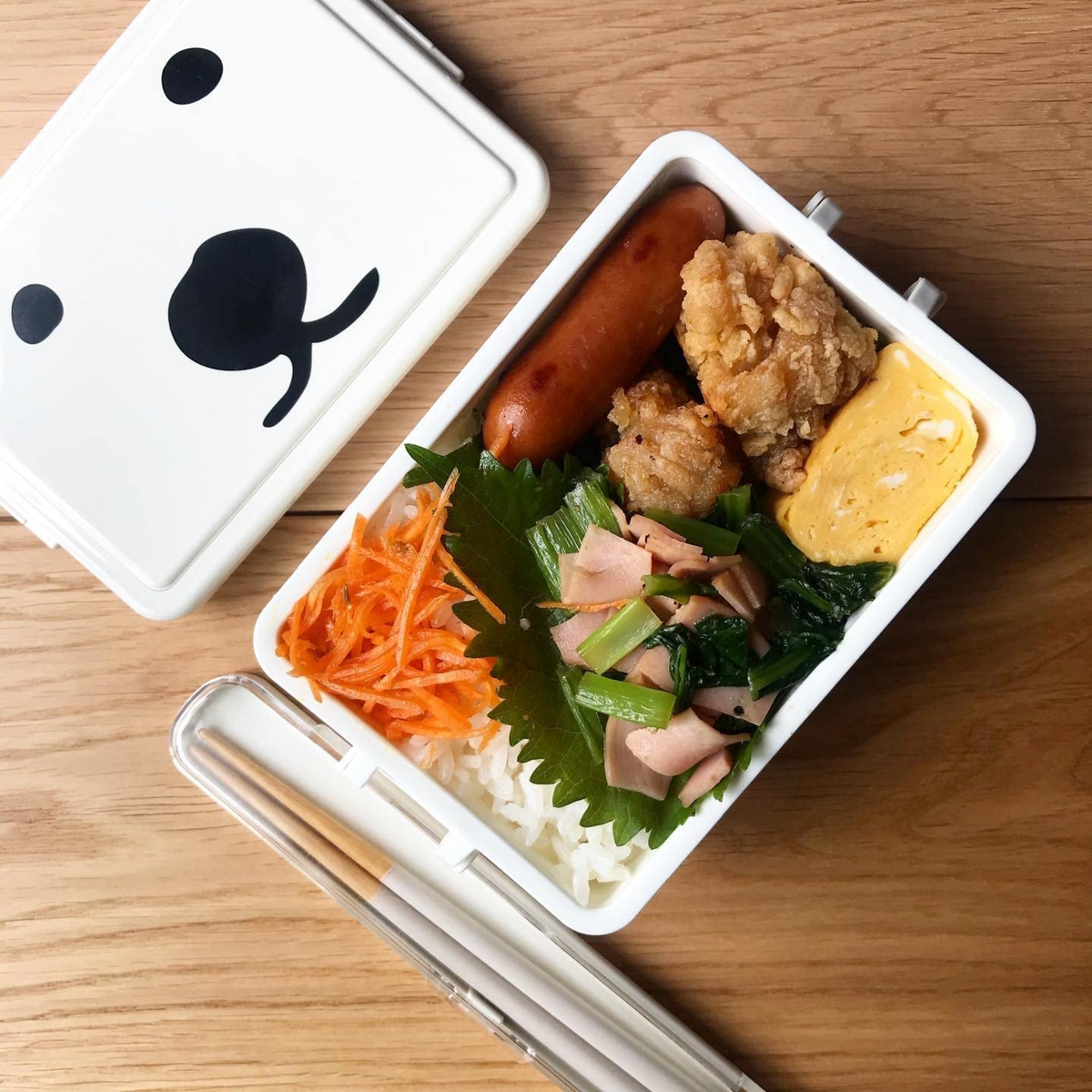 Overhead view of Gel-Cooma Bento Box Medium 400ml open on a wooden table, filled with white rice, shiso leaves, shredded carrots, sautéed spinach with ham, fried chicken, sausage, and tamagoyaki, with the white bear-face lid and a clear chopstick case placed beside it