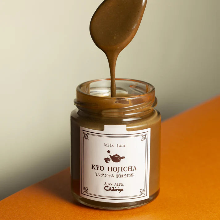 Chikiriya Kyo Hojicha Milk Jam dripping from a spoon into an open glass jar, showing the smooth, creamy brown texture, on an orange surface