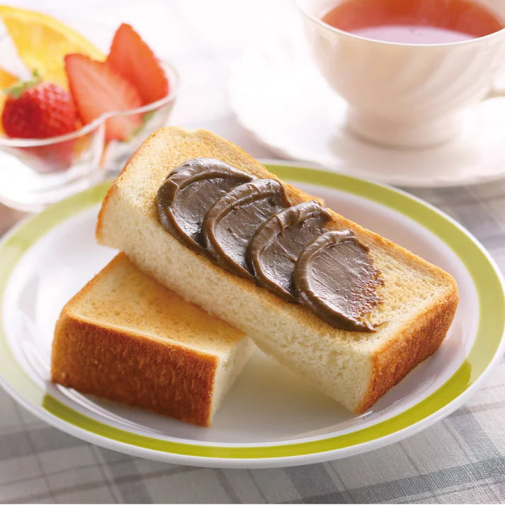 Chikiriya Kyo Hojicha Milk Jam spread in thick stripes on a slice of white toast on a green-rimmed plate, served with fresh strawberries, orange slices, and a cup of tea