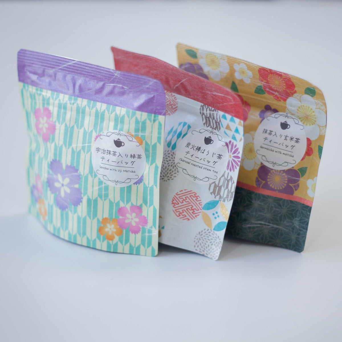 Three Chikiriya tea pouches from the Wafu Green Tea Gift Set displayed at an angle – green floral Uji Matcha Green Tea in front, white geometric Charcoal Roasted Hojicha in center, yellow floral Genmaicha with Matcha at back, on white background