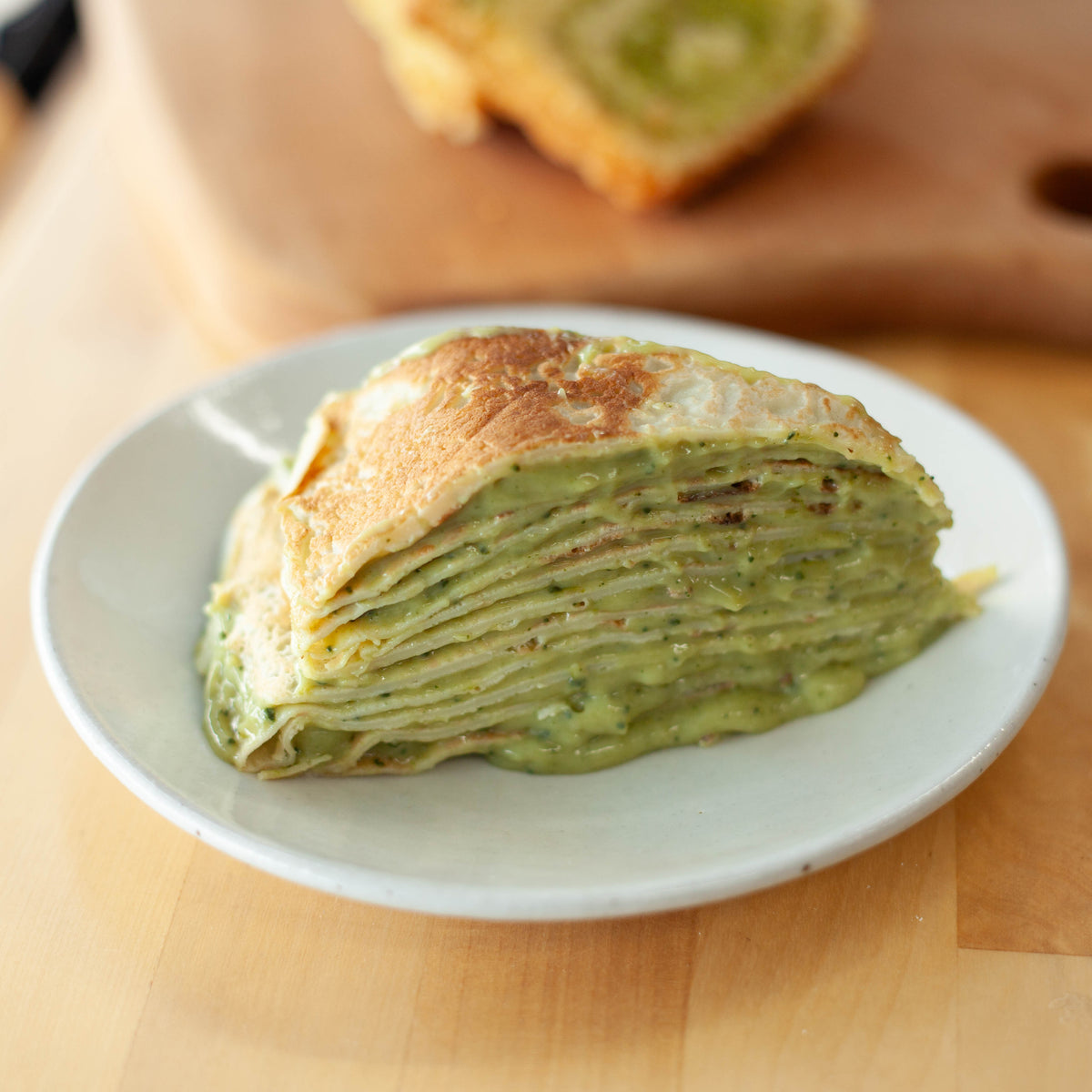 Slice of matcha mille crepe cake made with Chikiriya Shizuoka Matcha Powder – layers of green crepe and matcha cream on a white plate, wooden surface background
