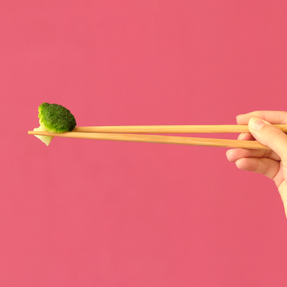 Hand using long bamboo chopsticks to hold a broccoli floret against a pink background
