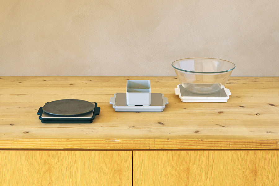 Three Gel-Cool Rapid Cooling Plates in black, grey, and white displayed on a wooden shelf – black plate with a round ceramic plate on top, grey plate with a two-compartment tray and glass bowl, and white plate with a glass mixing bowl, showing versatile cooling uses