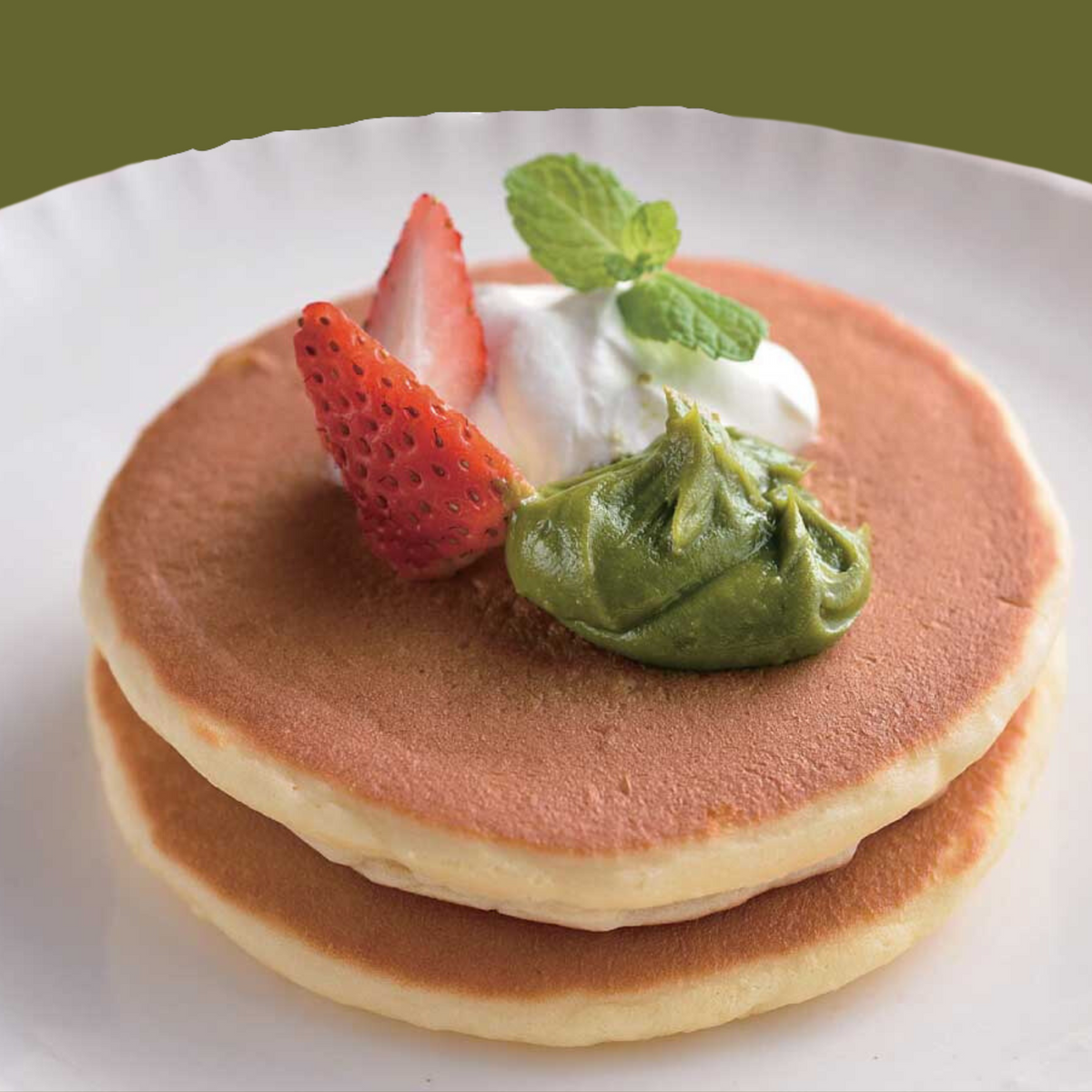 Stack of two fluffy pancakes topped with a dollop of Chikiriya Uji Matcha Milk Jam, whipped cream, and fresh strawberry slices, on a white plate with a green background