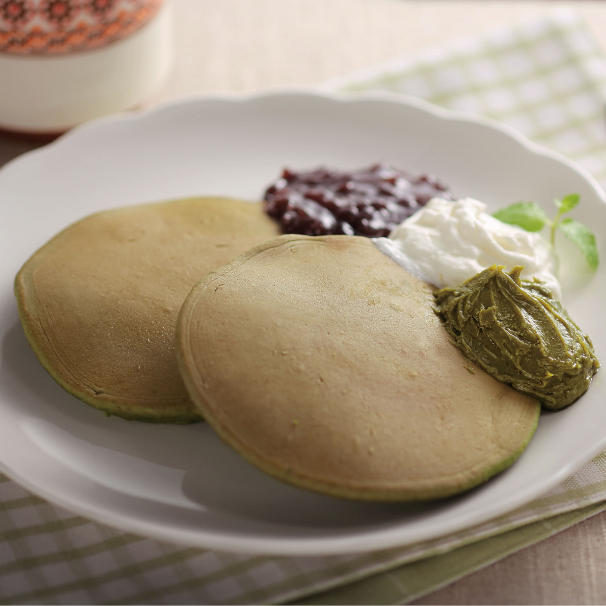 Two green matcha-flavored pancakes on a white flower-shaped plate, topped with Chikiriya Uji Matcha Milk Jam, whipped cream, and red bean paste, on a wooden table with a green cloth