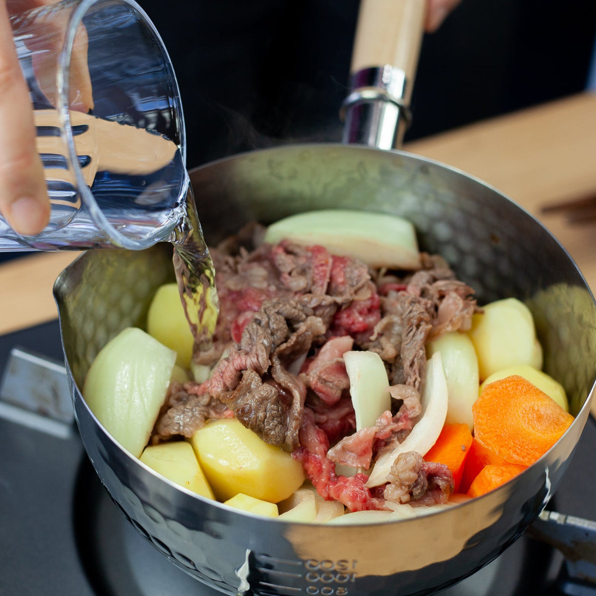 Pouring water into YukiHira Nabe Pot filled with beef, potatoes, carrots and onions for nikujaga stew on induction ho