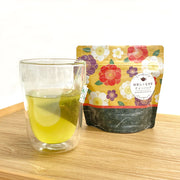 Chikiriya Wafu Genmaicha with Matcha tea bag steeping in a double-walled glass mug showing vivid yellow-green tea, beside the yellow floral Japanese-pattern resealable pouch, on a wooden surface