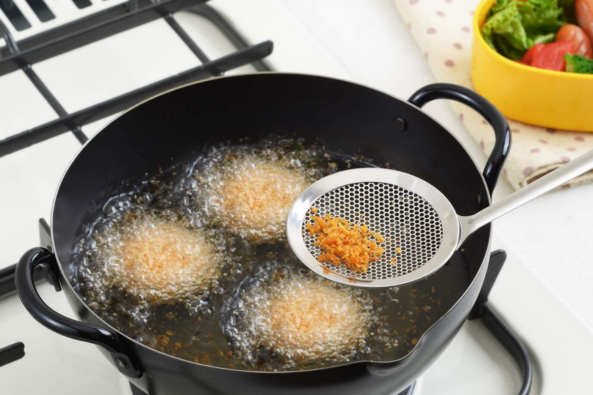 Perforated Oil Skimmer scooping fried breadcrumb bits from hot oil while korokke fry in black iron tempura pot, side angle view