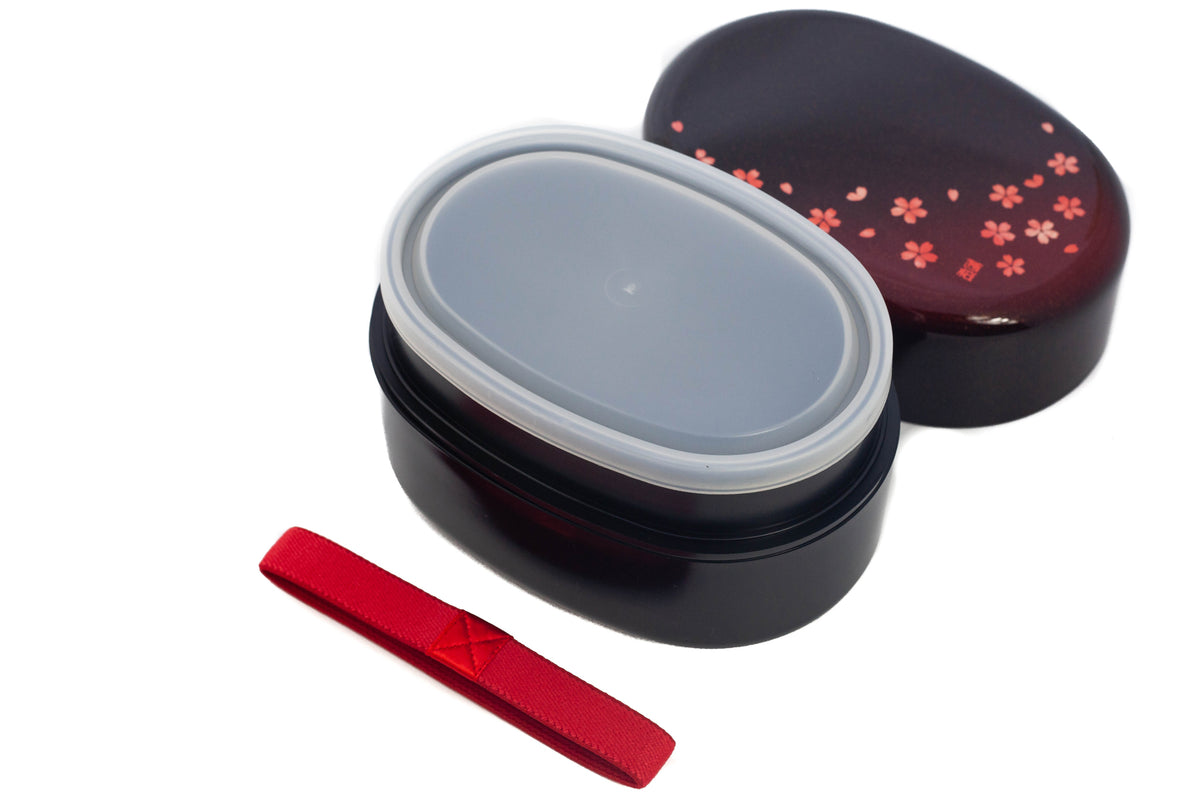 Hakoya Sakura Petals Oval Bento Box closed, black to burgundy lacquer with scattered pink cherry blossom pattern, angled view on grey background