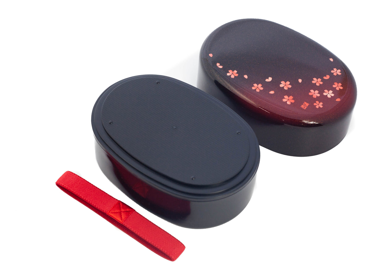 Hakoya Sakura Petals Oval Bento Box closed, black to burgundy lacquer with scattered pink cherry blossom pattern, angled view on grey background