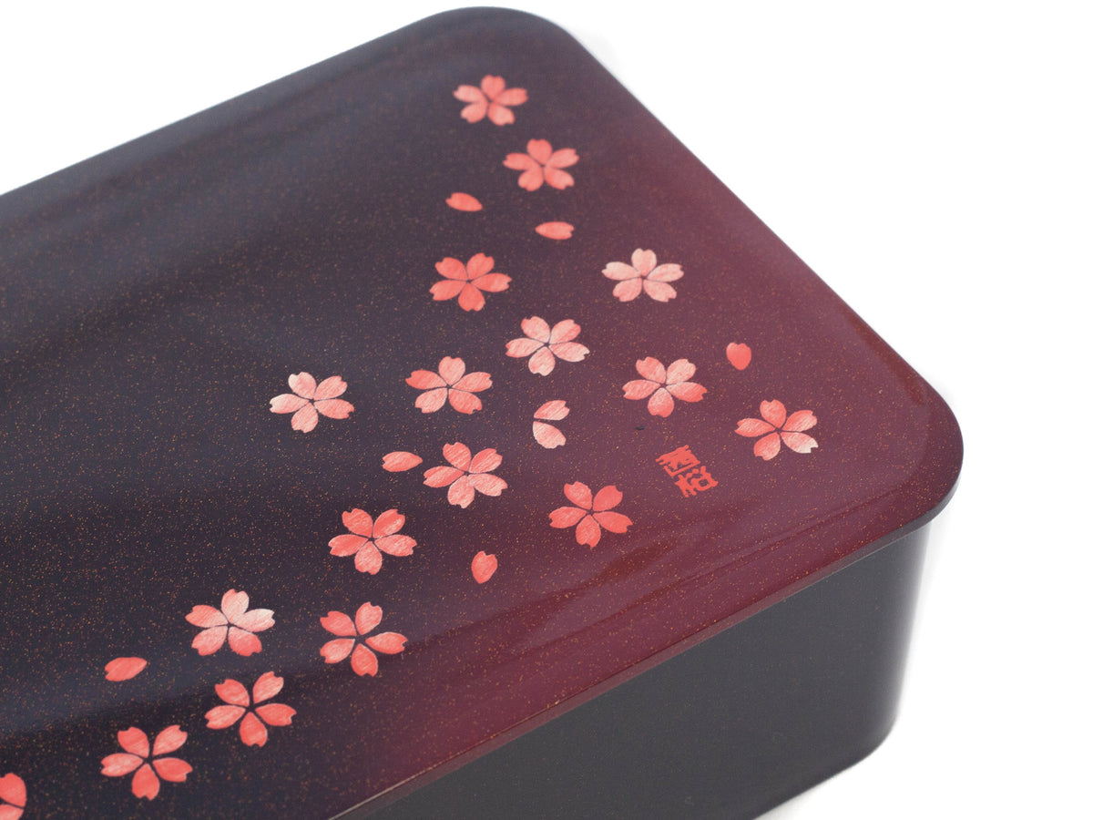 Hakoya Sakura Petals Bento Box closed, black to burgundy gradient lacquer lid with scattered pink cherry blossom pattern, angled view on grey background